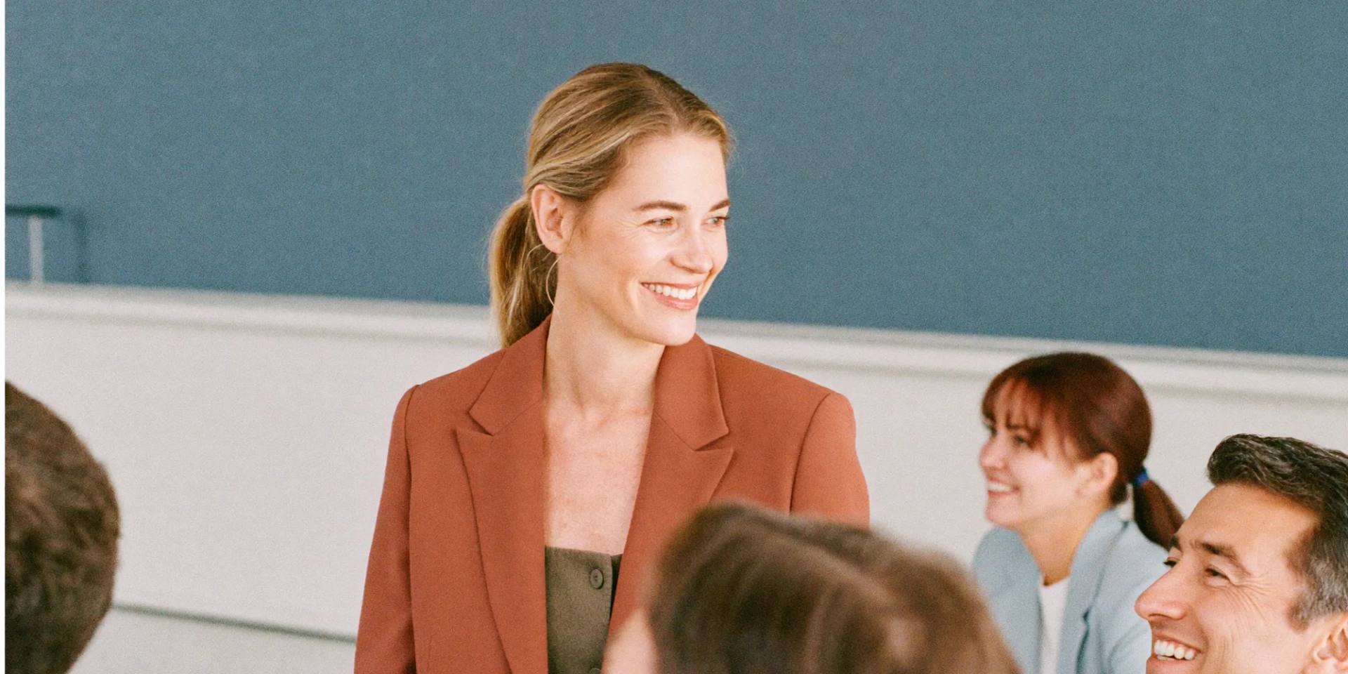 A smiling woman in a rust-red blazer stands in front of a small group listening to her.